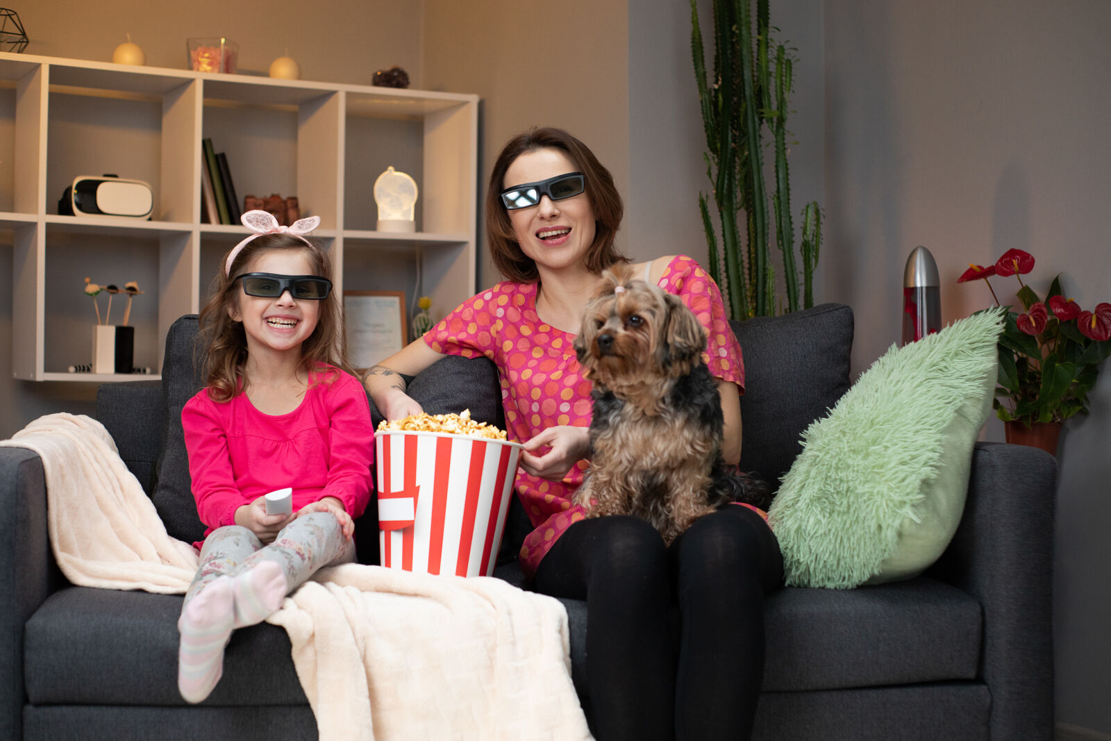 Mama z psem na kolanach i córką obok w okularach 3D oglądają film jedząc popcorn.